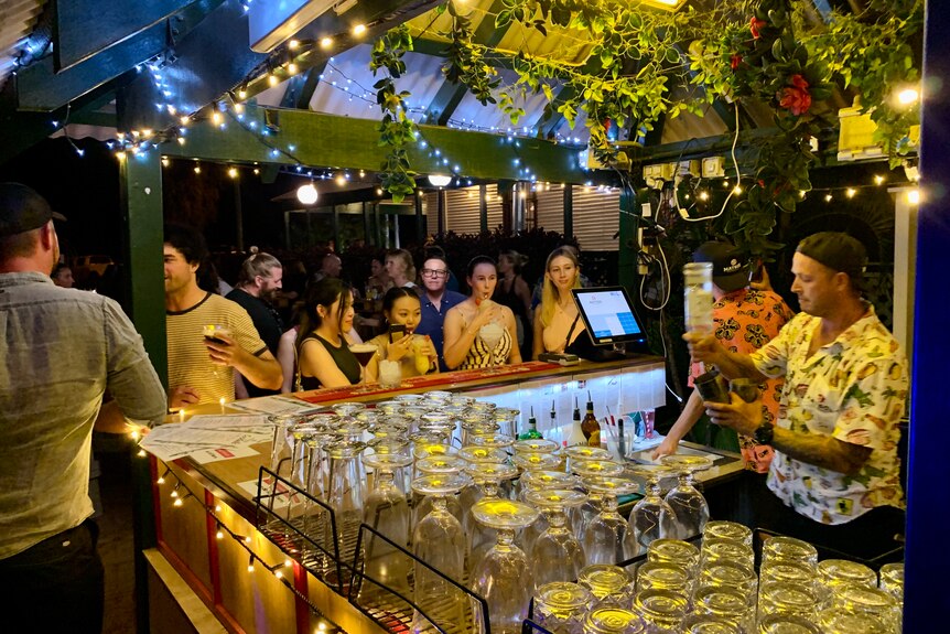 Young people line up at an outdoor bar festooned with pot plants and fairy lights
