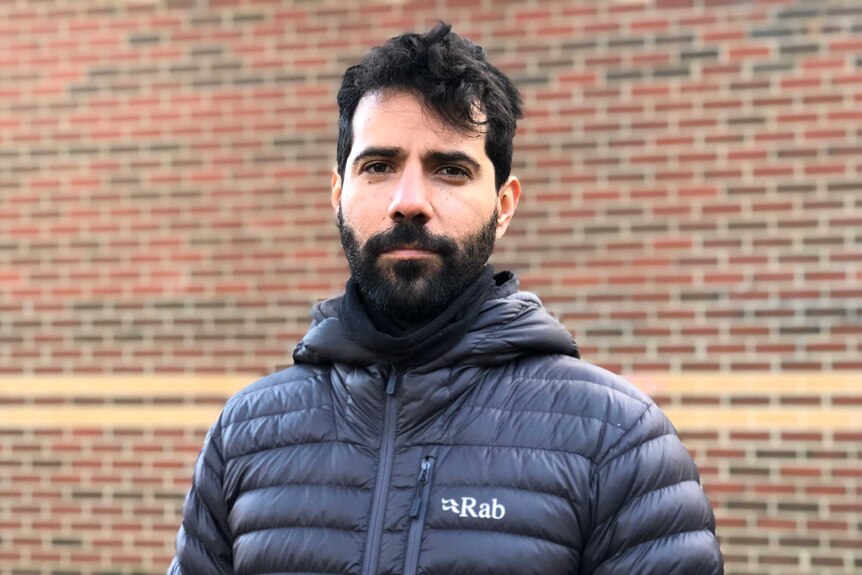A man with dark hair and a beard wears a black puffer jacket and stands outside in front of a brick wall.