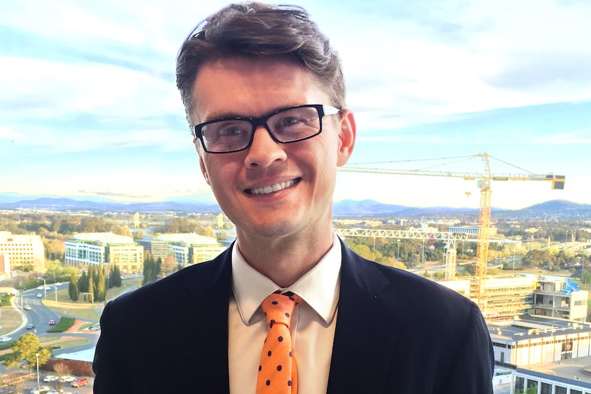 A man with brown wavy hair and black rectangular glasses, wearing white shirt, orange tie, black suit smiles and Canberra behind