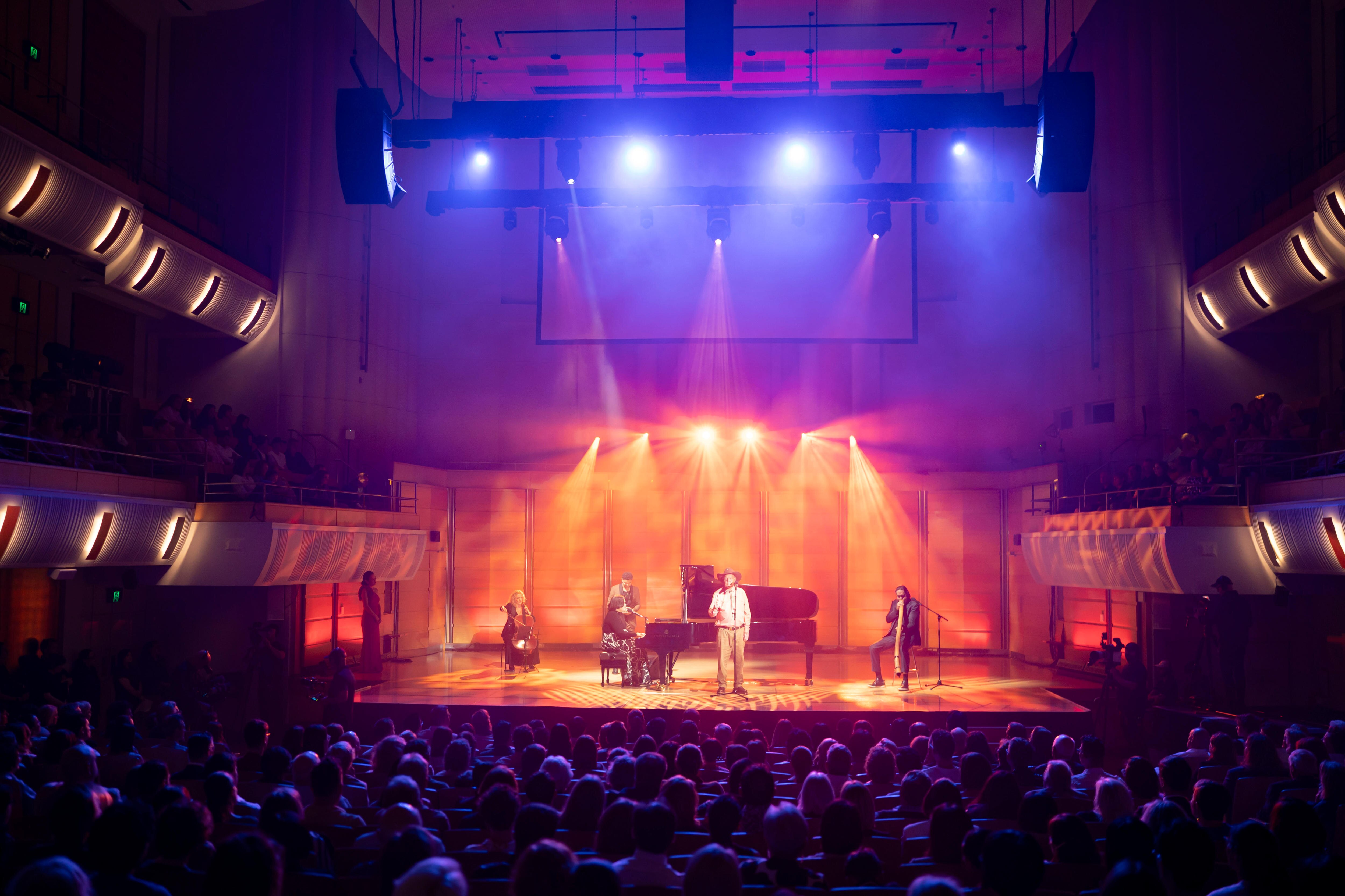 A large stage lit up in orange and blue lights, with Peter on the microphone, Rosie at the piano, a cellist and Barton on yidaki