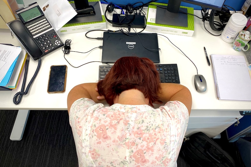 A woman sitting at office work station with her head on the desk.
