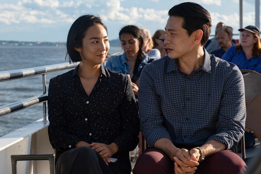 Greta Lee, an Asian woman in a black blouse, and Teo Yoo, an Asian man in a plaid shirt, sit together on a ferry deck.