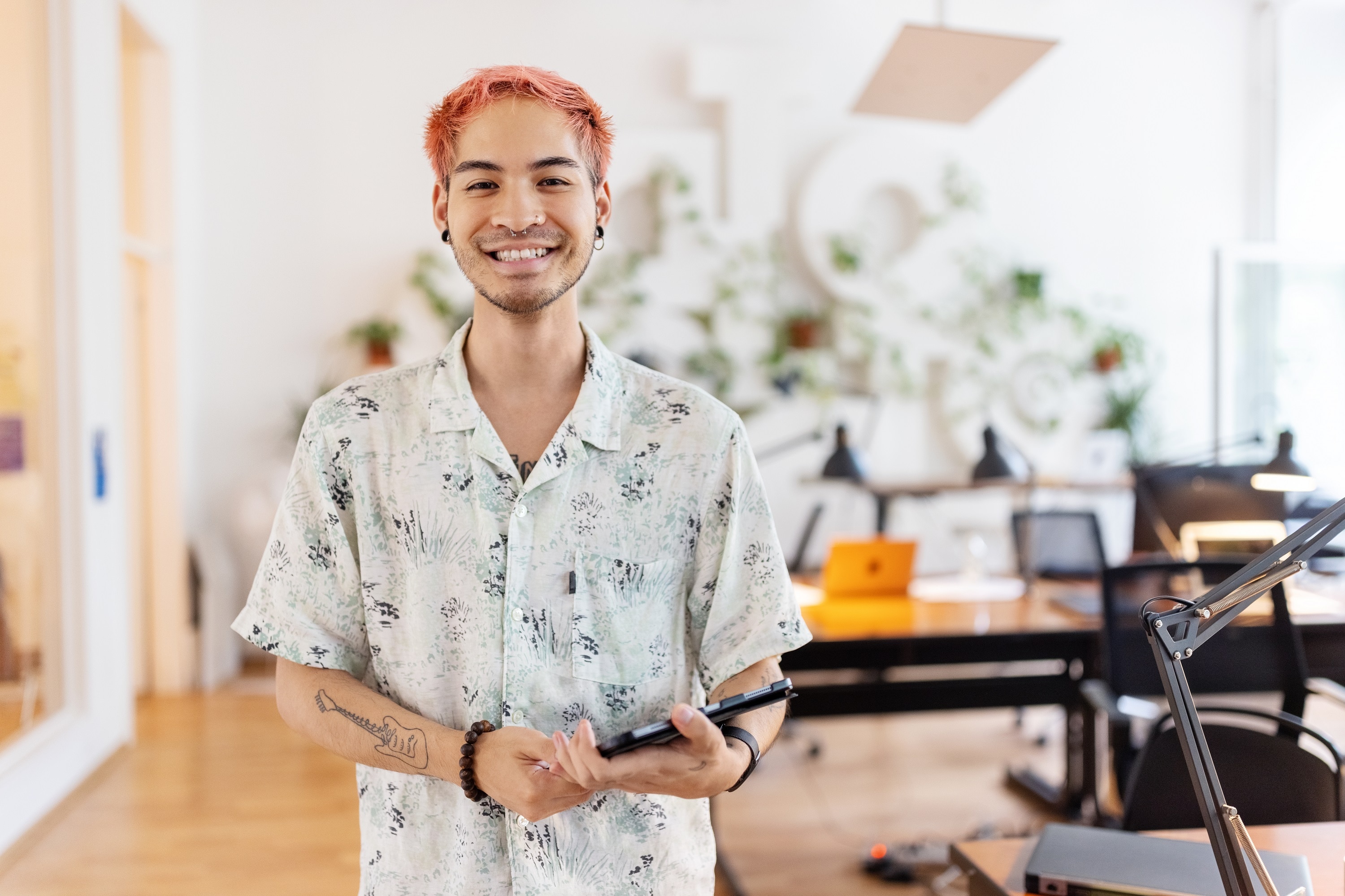A young Asian man with pink hair and piercings stands in an office holding a tablet