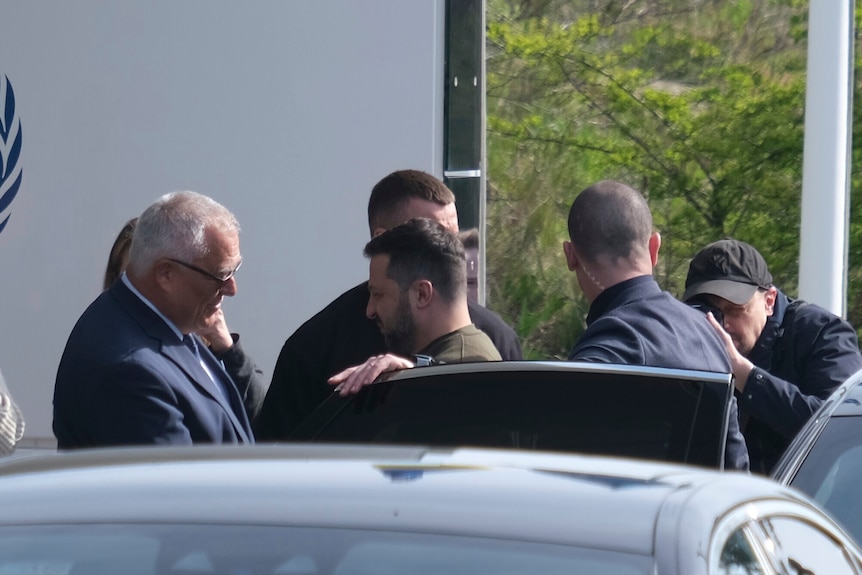 A middle-aged man with short dark hair in a khaki tracksuit top exits a car to greet a man with white hair in a suit.