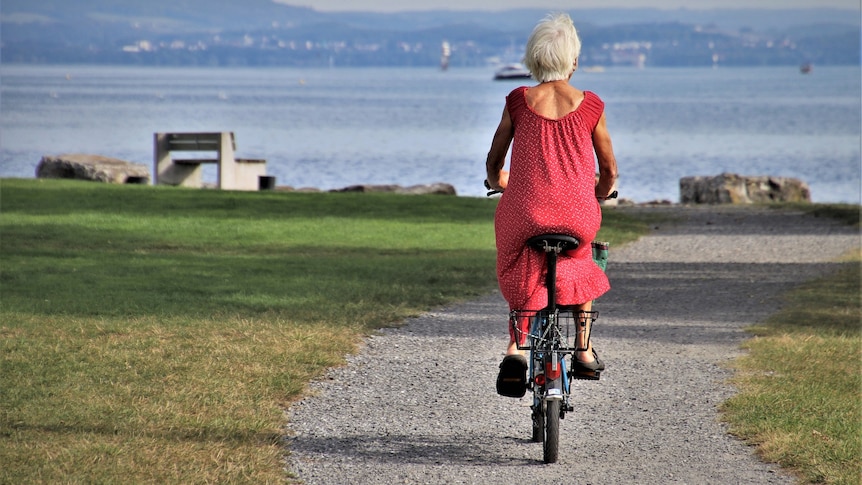 The back of a woman with grey hair and pink dress riding a bike on a path towards the sea.