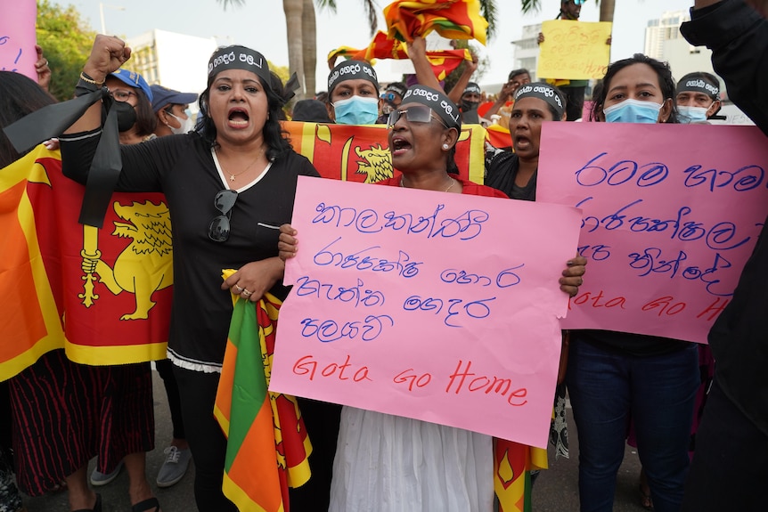 Three women wearing face masks at the front of a protest crowd hold up pink cardboard signs saying "Gota Go Home"