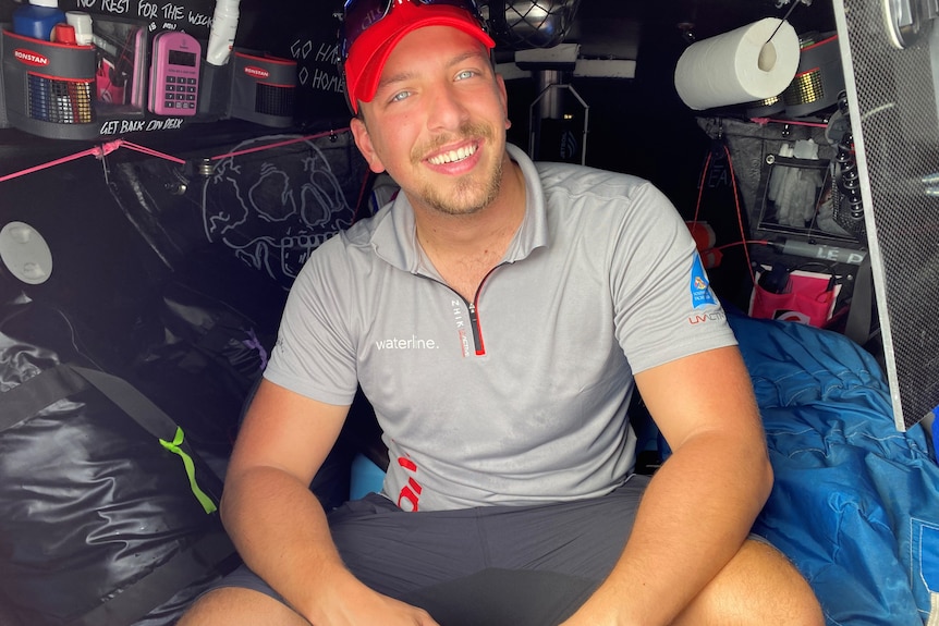 Man wearing red cap and gray shirt sitting in cabin of small yacht with radio equipment in background.