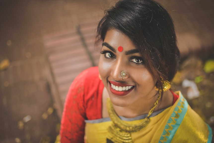 A woman smiling with a red bindi on her forehead and a gold nose stud in her nostril