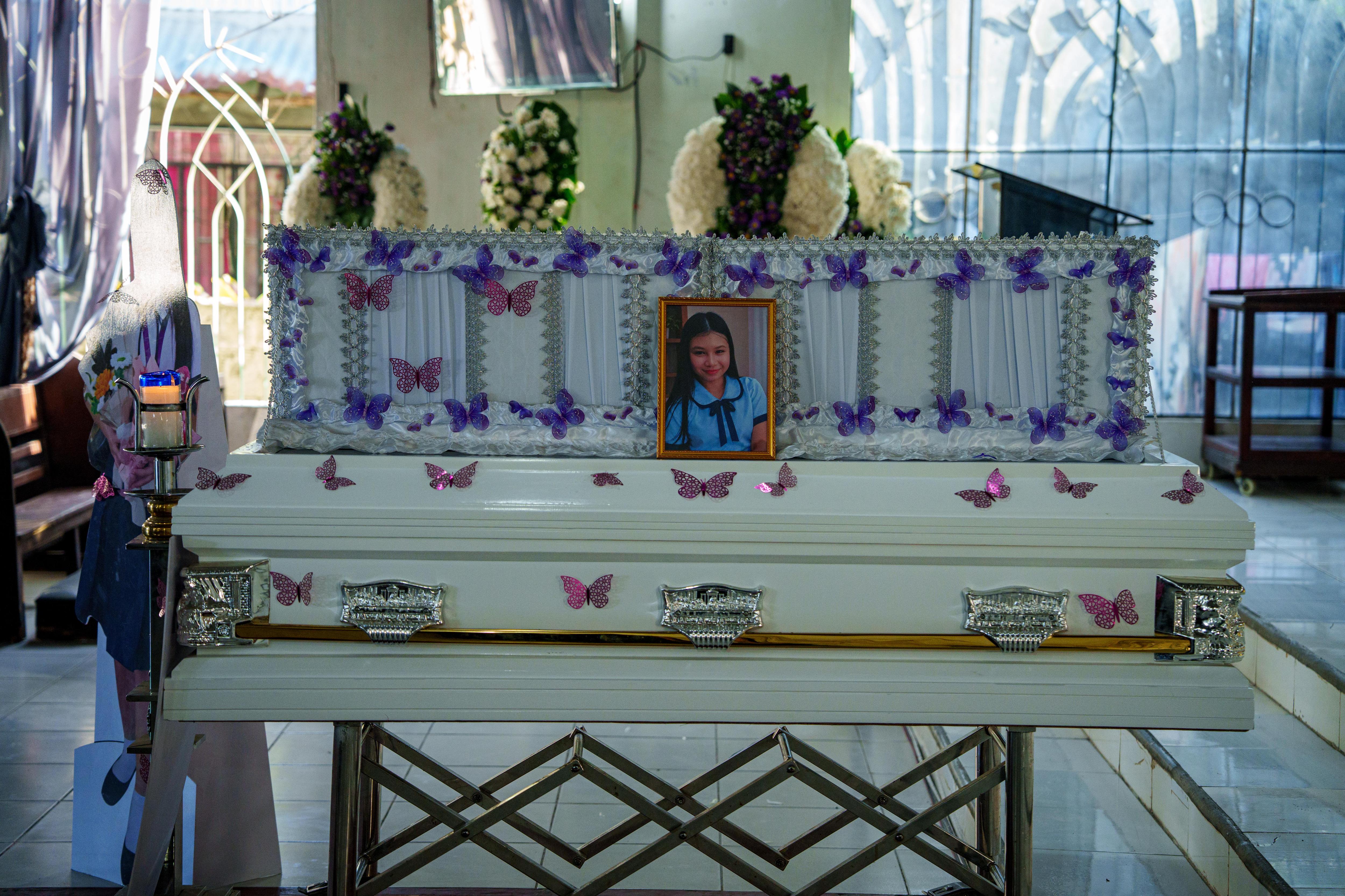 A white coffin decorated in purple butterflies rests in a church with a framed photo of a school girl on top.