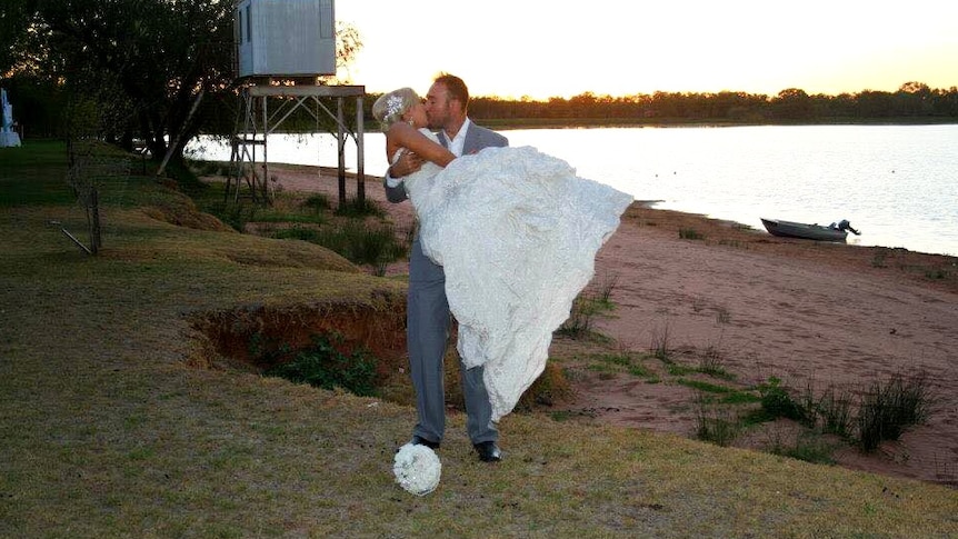 A man in a suit kisses a woman in a wedding dress, next to a lake.