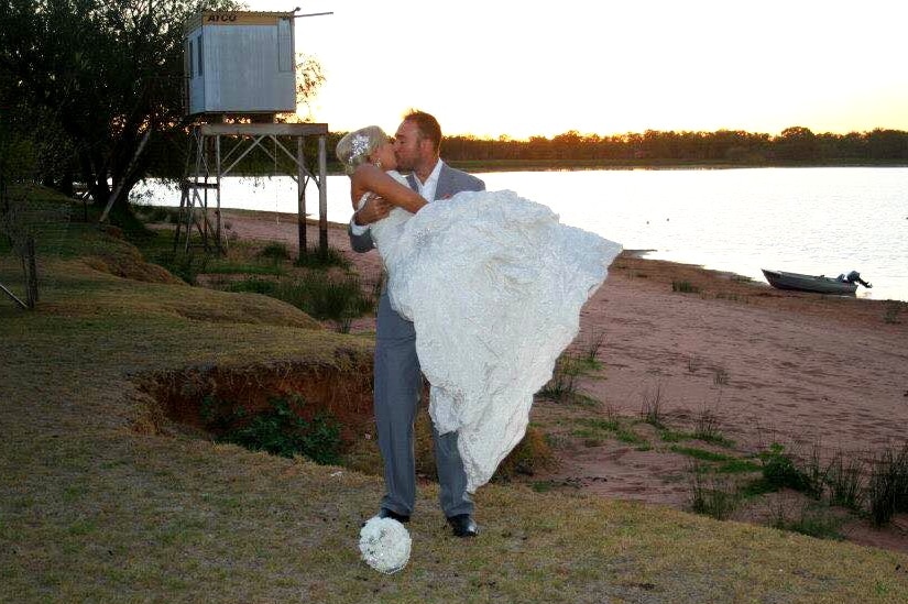 A man in a suit kisses a woman in a wedding dress, next to a lake.