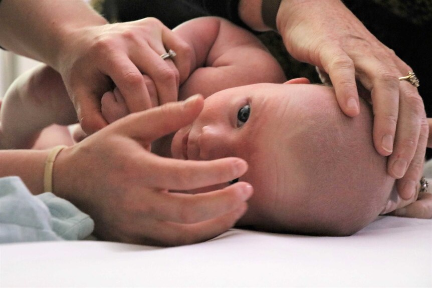 A baby lies on a table, women's hands on his head and holding his arm.
