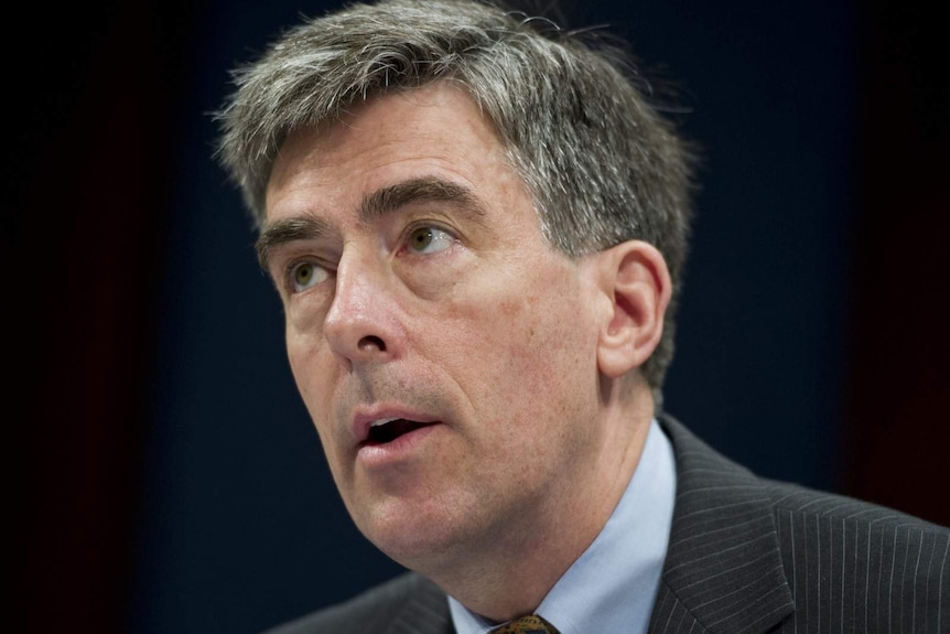 Former deputy director of the US NSA Chris Inglis looks into the camera standing in front of two flags.