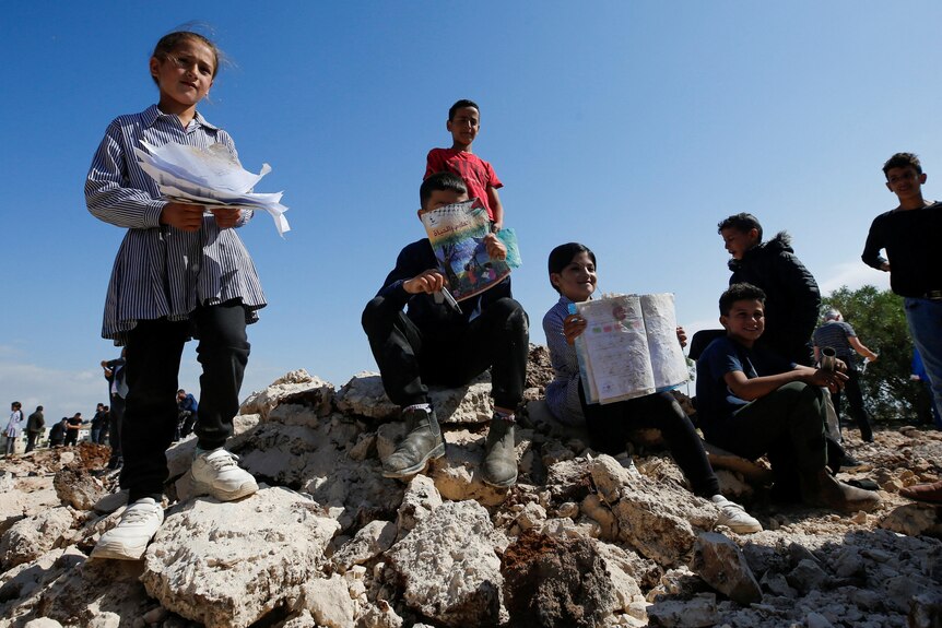 Children stand atop rubble with textbooks and papers in their hands.