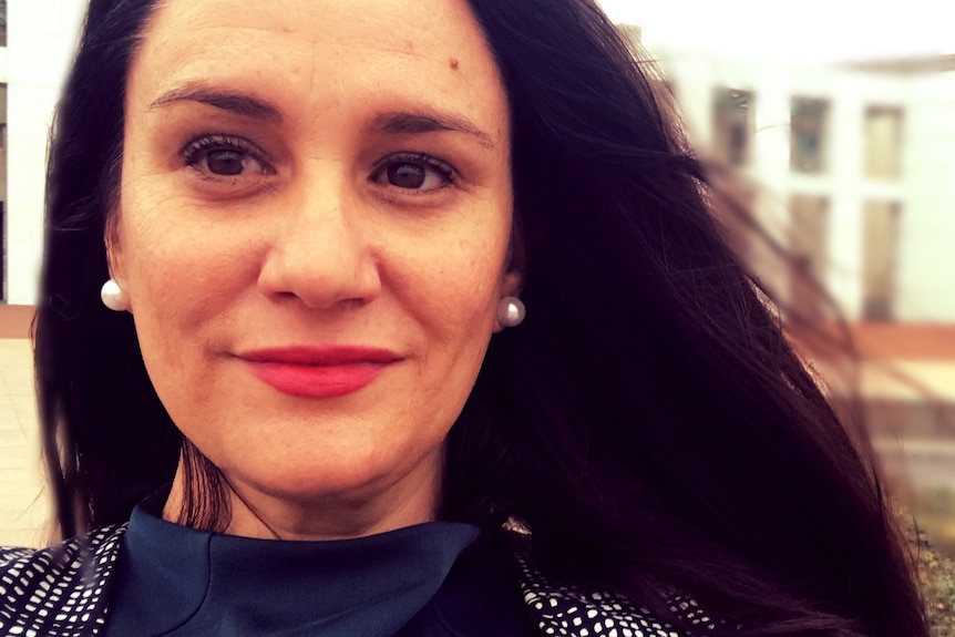 A smiling woman stands in front of parliament house, dark long hair, wears pears, red lispstick.