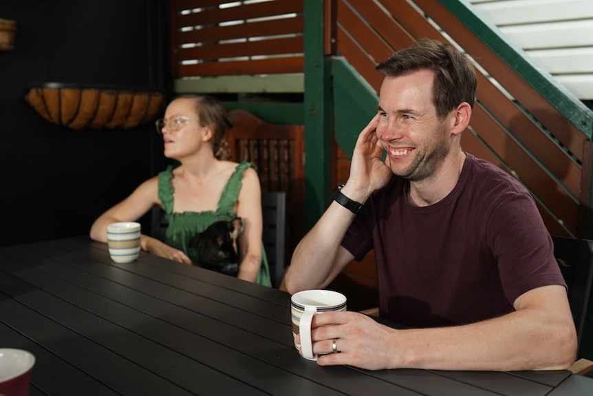 A man and woman sit at an outdoor dining table with mugs.