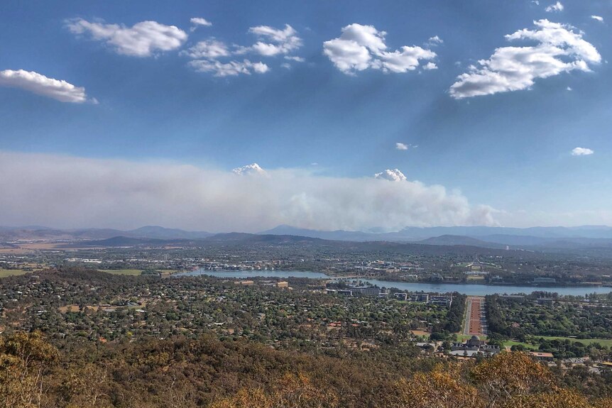 Canberra is sprawled out below, a large amount of smoke billowing out in the distance, blue sky and clouds above.