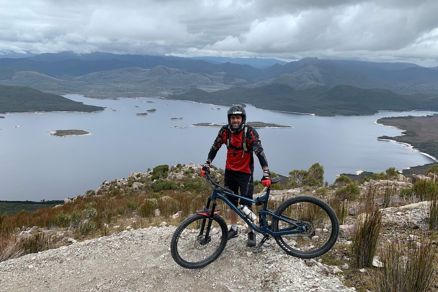 A man with full face helmet and bike poses in front of a huge lake view from mountain top.