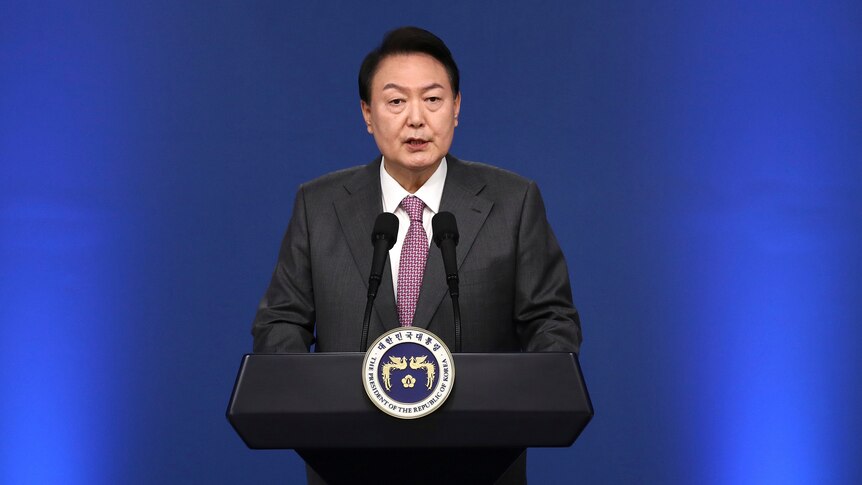 An older-looking Korean man in a suit speaks into a microphone at a podium in front of a deep blue background.