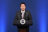 An older-looking Korean man in a suit speaks into a microphone at a podium in front of a deep blue background.