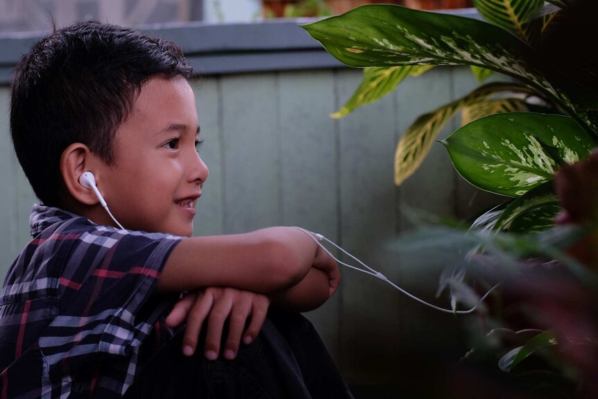 Young boy sits outside with headphones in his ears. He is smiling at something he's listening too.