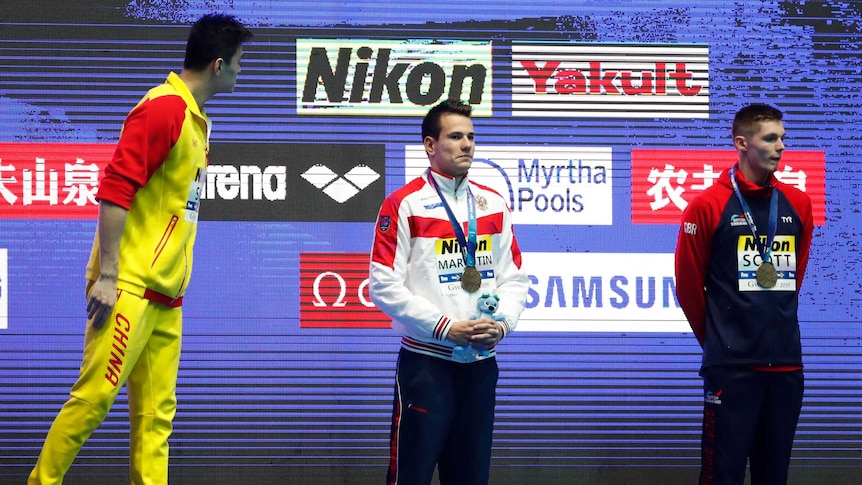Sun Yang, in yellow and red Chinese team uniform, turns towards two other swimmers at the podium