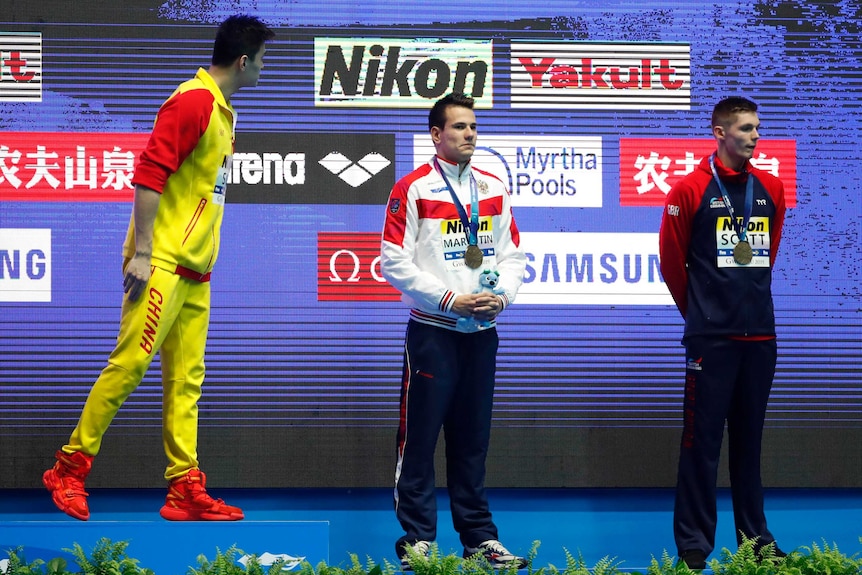 Sun Yang, in yellow and red Chinese team uniform, turns towards two other swimmers at the podium