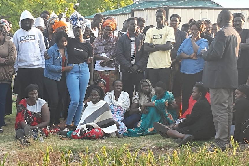 Family and friends of Aguer Akec stand and sit as a group on grass mourning the death of Aguer Akec.