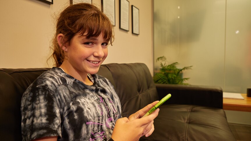 A teenage girl holding a smartphone sits on a couch and smiles at camera 