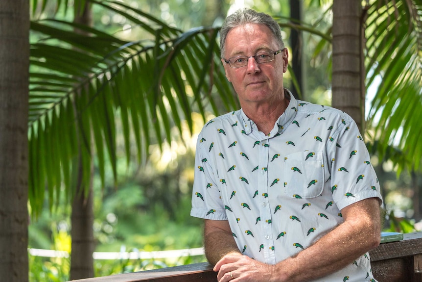 A man wearing a button up shirt with parrots printed on it stands in front of palm trees