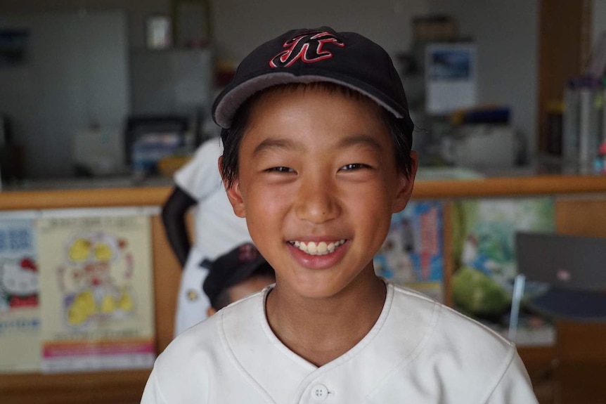 A young Japanese boy wearing a baseball cap smiles as he looks into the camera.