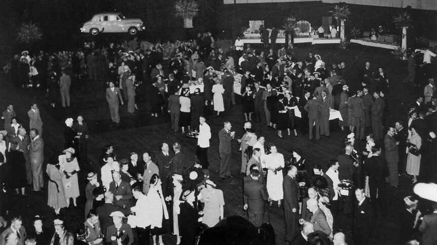 A black and white photo from 1948 showing crowds of people in a hall with an old Holden on display