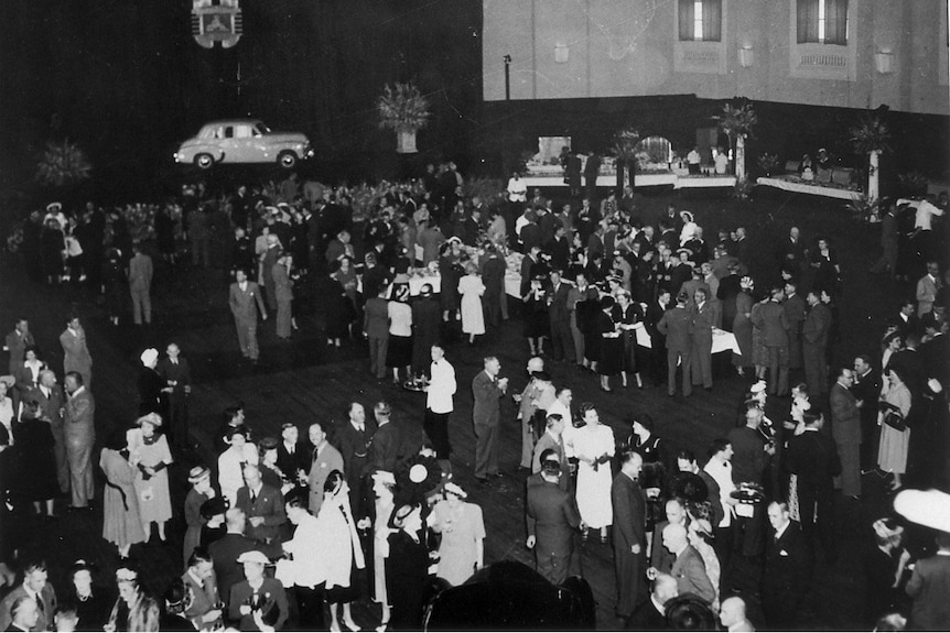 A black and white photo from 1948 showing crowds of people in a hall with an old Holden on display