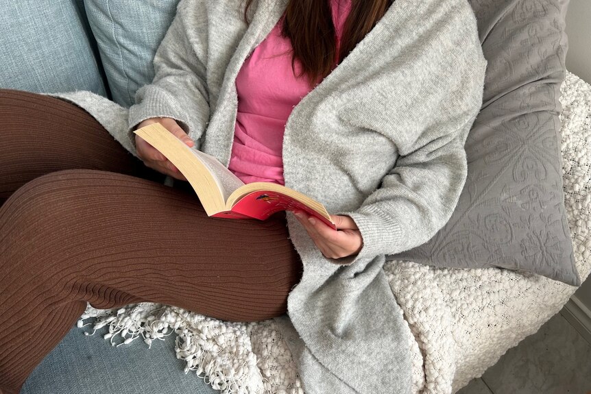 A person sits on a couch reading a book, with the image deliberately framed to obscure her face. 