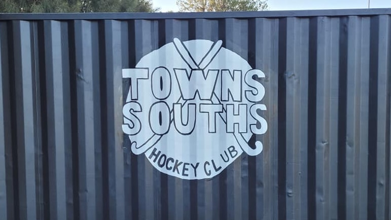 A sky blue circle with two crossed hockey and words Towns Souths Hockey Club logo painted on a darker blue shipping container.