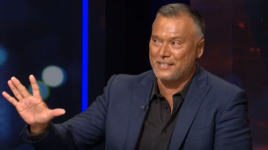 Australian journalist Stan Grant raises his hand to make a point, while half smiling.