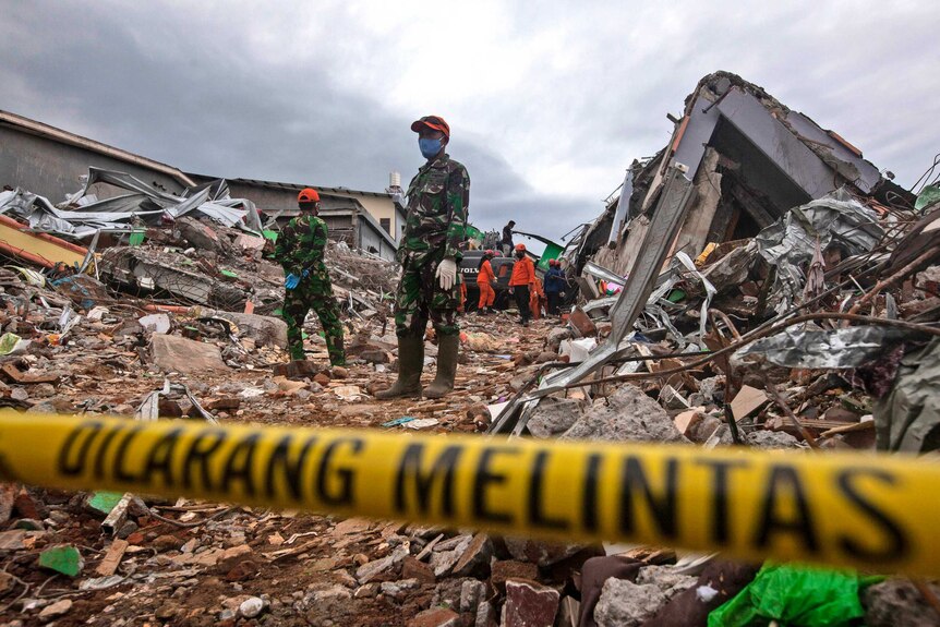 Soldiers stand guard in front of yellow tape as rescuers search for survivors in a pile of rubble behind them.