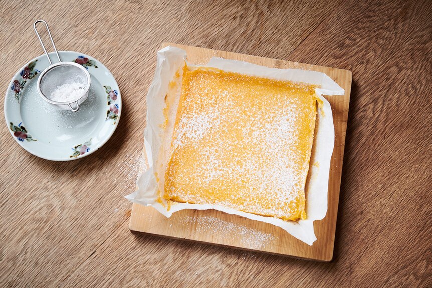 A square of lemon bars dusted with icing sugar sitting on a cutting board before being sliced.
