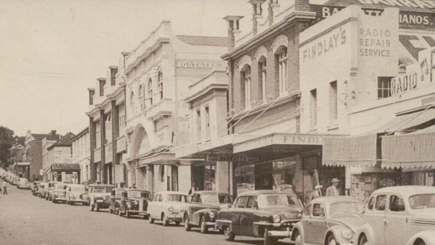 An old photo of a row of buildings with cars parked in front.
