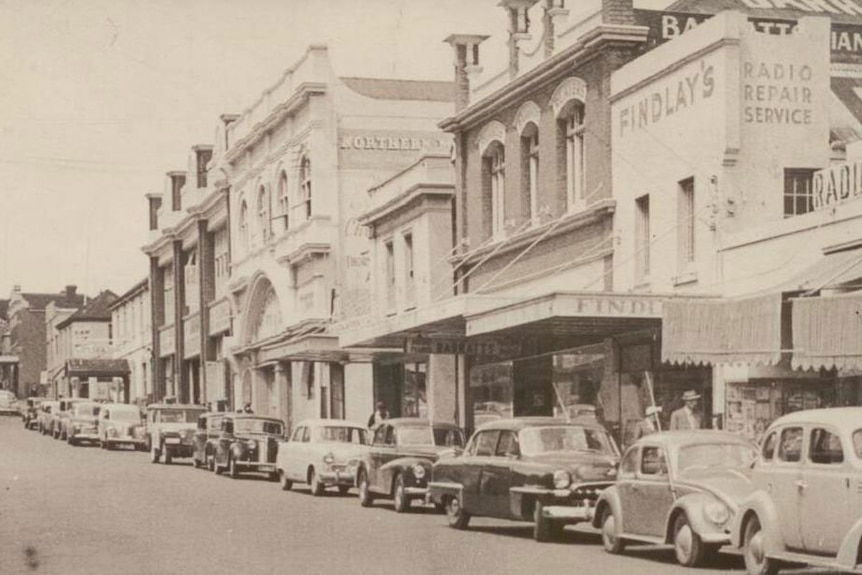 An old photo of a row of buildings with cars parked in front.