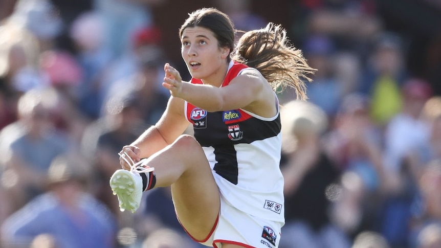 A young AFLW player kicks a ball and looks towards it with a crowd behind her in the stands.
