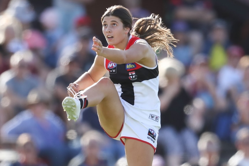 A young AFLW player kicks a ball and looks towards it with a crowd behind her in the stands.