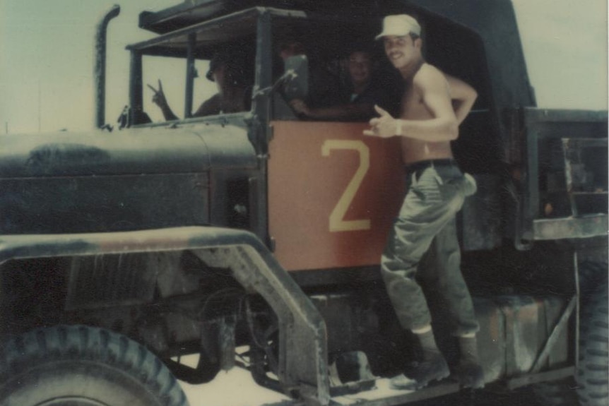 A shirtless soldier stands on the running board of a truck to pose for a photo with three other soldiers in the truck.