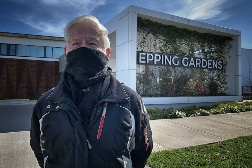 Man with grey hair using a scarf to cover his mouth and nose outside a building with a sign reading Epping Gardens.