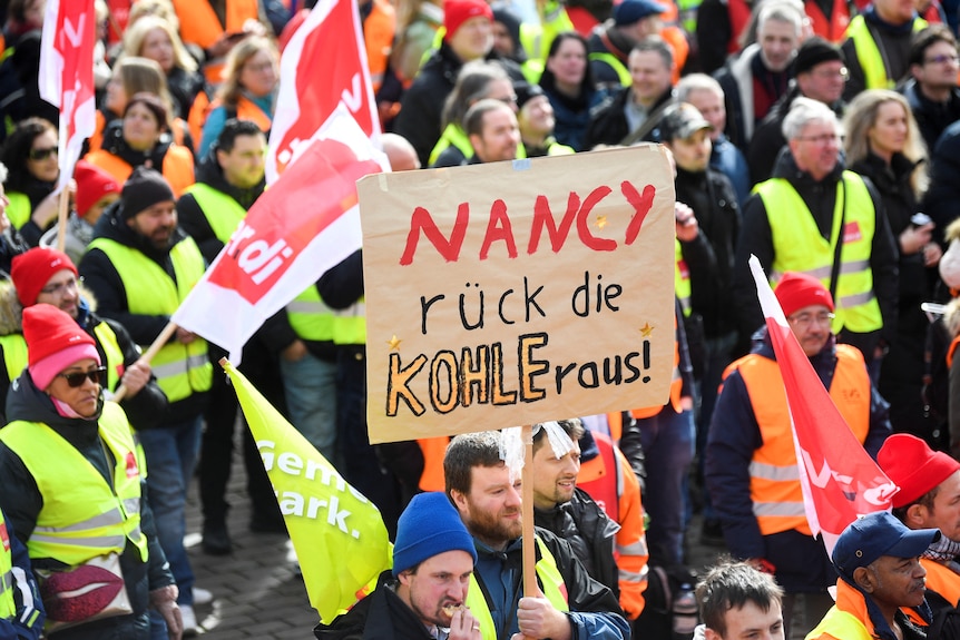 A group of people wearing high-vis vests while waving flags. One of them holds up a sign that says 'Nancy rück die kohle raus!'.