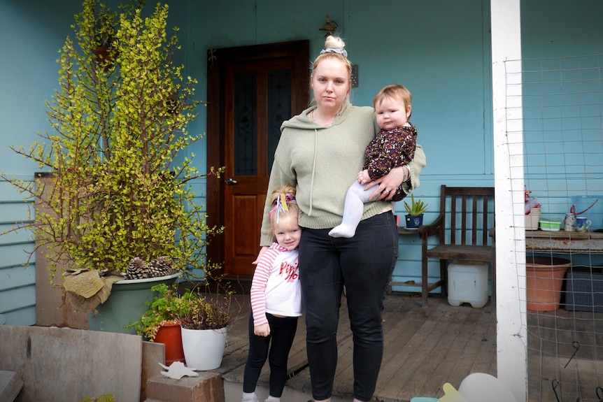 A blonde woman with her hair in a bun stands out the front of a house with two small children.