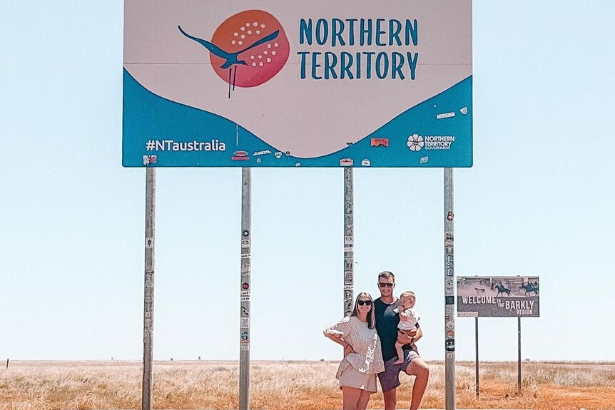 A woman and a man and a baby pose for a photo under an outback desert sign reading 'Northern Territory'