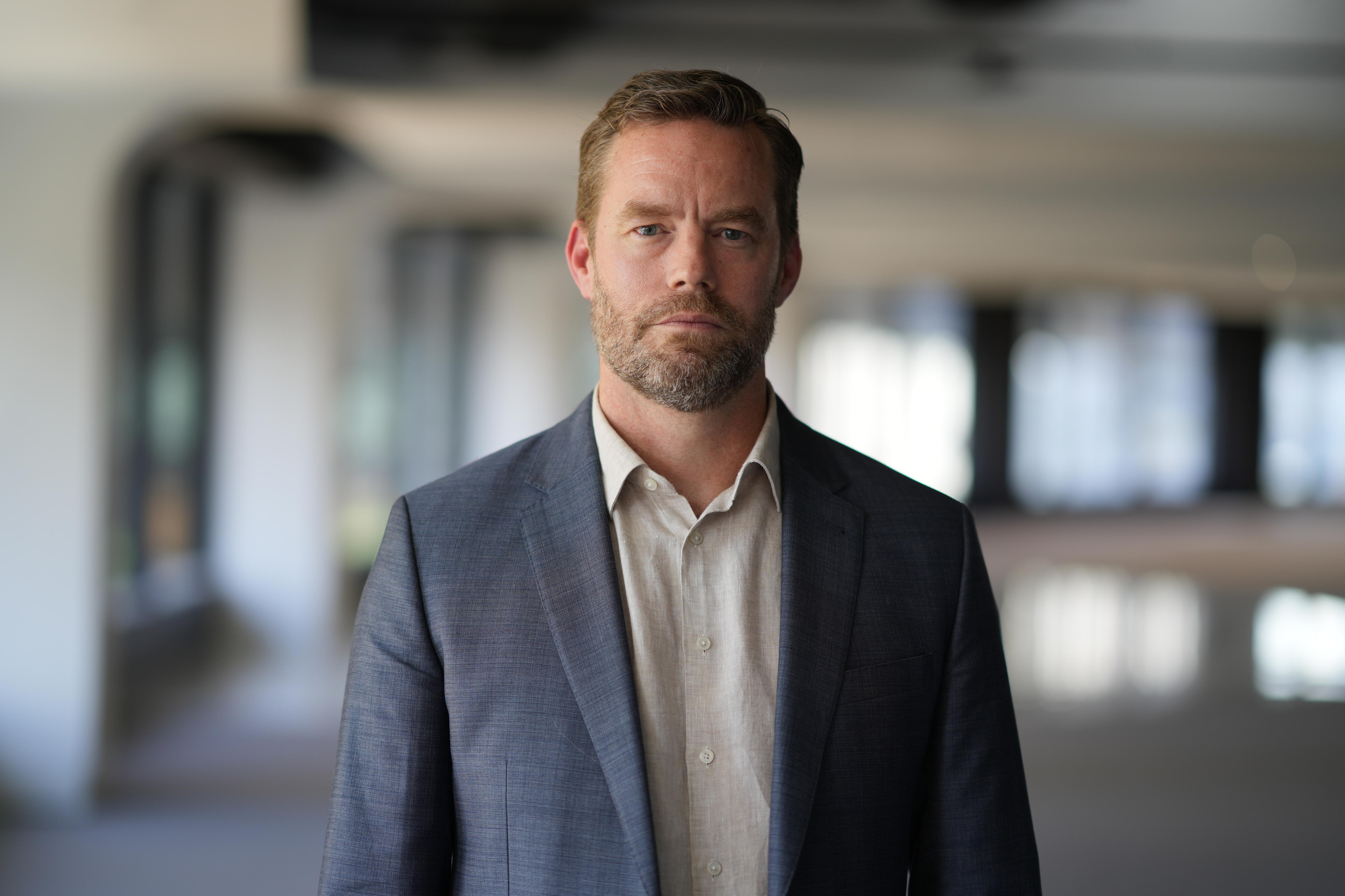 A man in suit jacket standing in a large empty room with windows. He is looking at the camera with a serious expression.