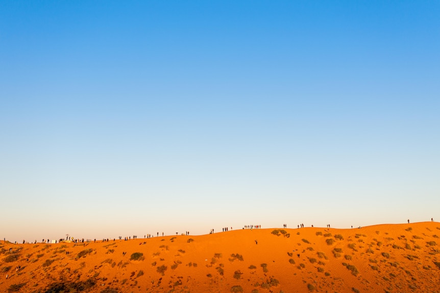 Small siilhouettes of people at the top of a giant sand dune at sunset