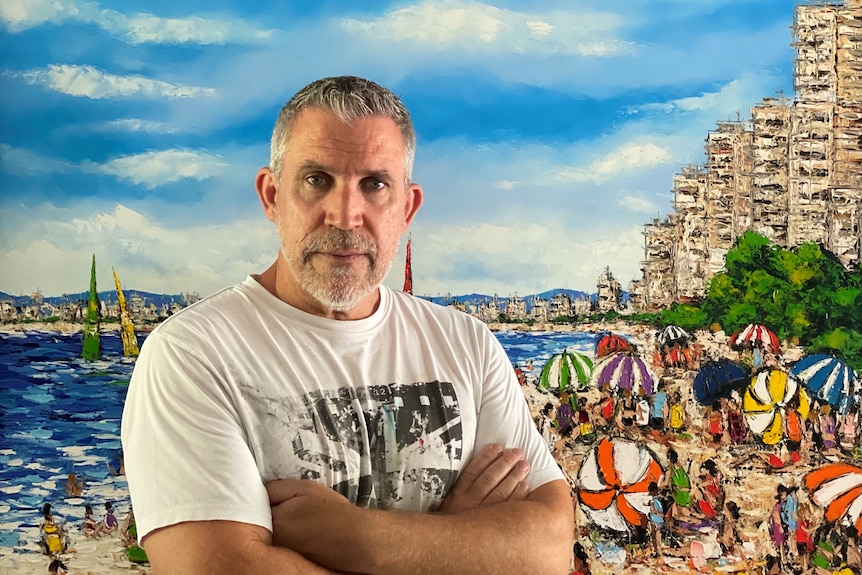 A man in his 60s in white tshirt with grey hair stands in front of a painting of a busy beach